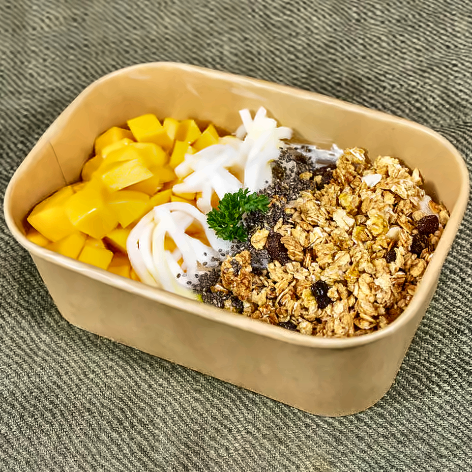 Acai bowl topped with mango, banana, coconut, chia seeds, granola, raisins, and parsley in a compostable container.