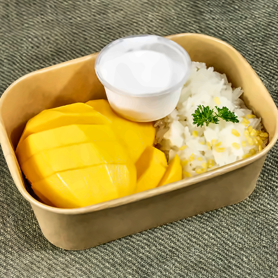 Takeout container holding sliced mango, sticky rice garnished with parsley, and a cup of creamy sauce.
