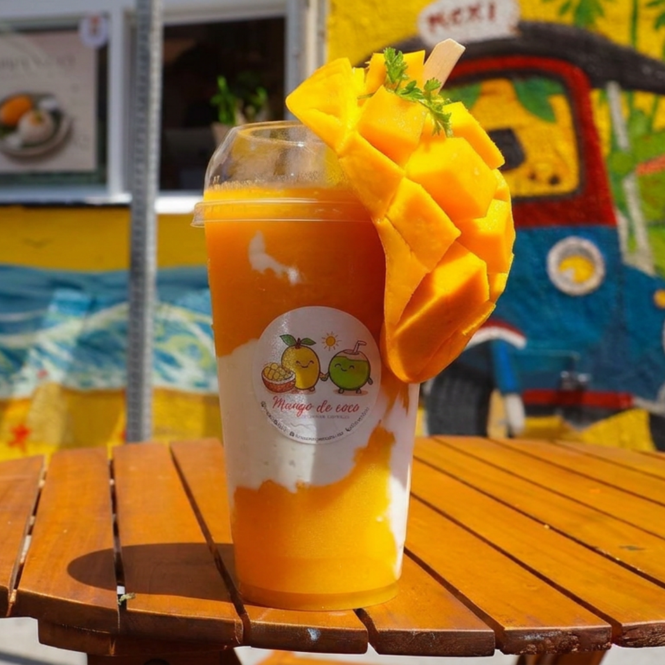 Mango smoothie topped with fresh mango chunks and herbs on a wooden table outside a colorful tropical restaurant.